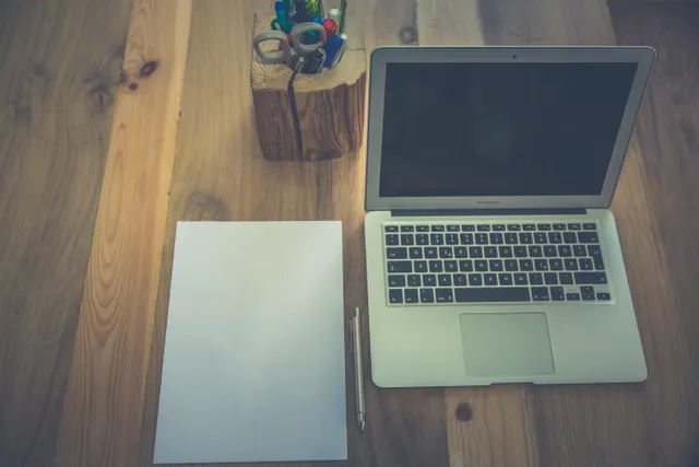 Laptop, blank paper, and pen on a wooden desk.